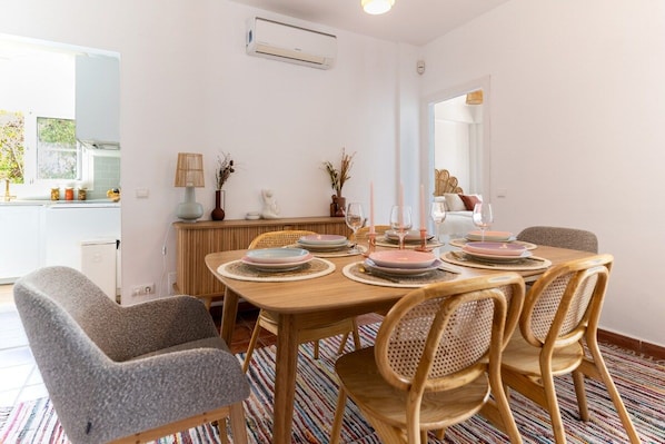 A dining area with a wooden table set for four, surrounded by wicker chairs, and a kitchen area with white cabinets and a window.