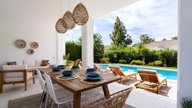 An outdoor dining area with a wooden table set for four, surrounded by wicker chairs, near a swimming pool with lounge chairs.