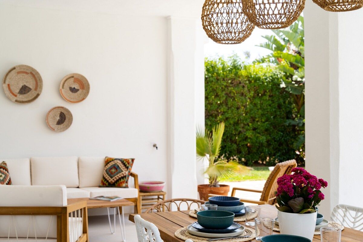 Une pièce comprenant une table à manger en bois, dressée avec des assiettes et des bols bleus, un canapé blanc agrémenté de coussins à motifs et des éléments décoratifs en fibres tressées accrochés aux murs.