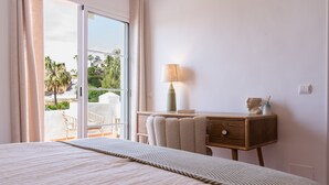 A bedroom with a large bed, a wooden nightstand, a lamp, and a view of the outdoors.