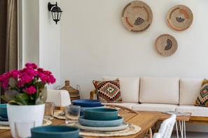 A dining area with a wooden table set with plates and glasses, a white sofa with patterned cushions, and wall-mounted woven art pieces.
