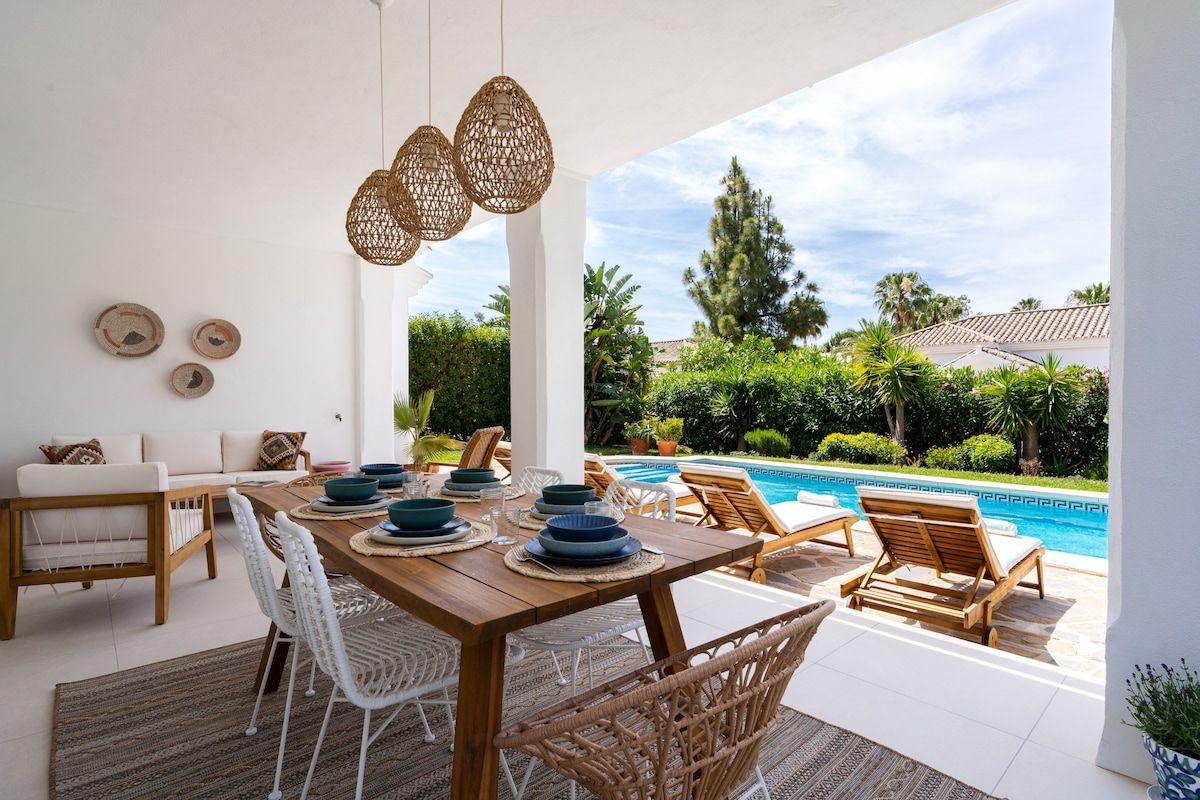Un espace de restauration en plein air avec une table en bois, des chaises blanches et des lampes en osier suspendues.