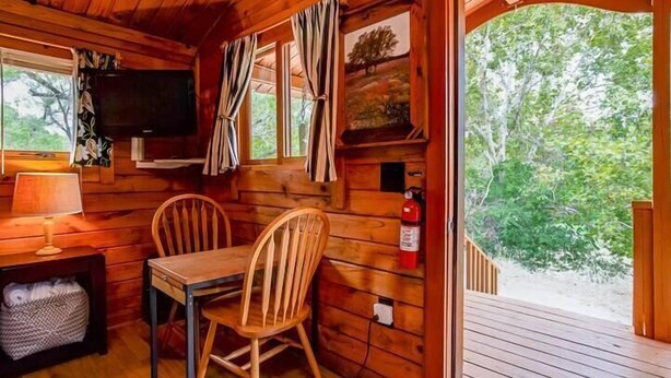 Interno di una casetta in legno con un tavolo, sedie, una televisione e una finestra con tende.