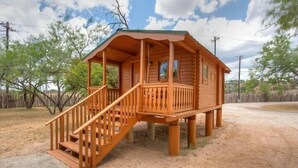 Una baita di legno con portico e scala, circondata da alberi e terreno sterrato.