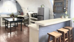 Et kompakt køkken med en central ø, komfur, mikroovn og køleskab. Der er barstole og et lille spisebord.