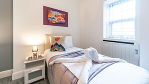 Une chambre à coucher avec un lit, une table de chevet avec une lampe, une fenêtre avec des stores et un tableau coloré au mur.