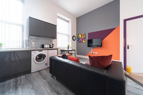 A compact living space with a kitchen, a washing machine, a sofa, a television, and a window with blinds.