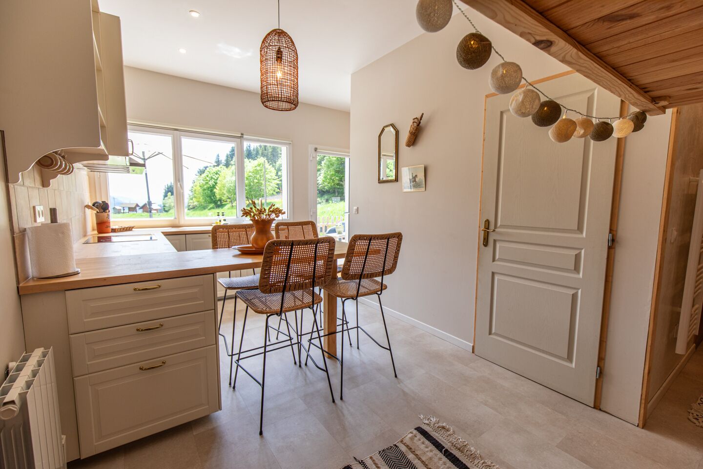 Une cuisine avec un coin repas, des meubles blancs, des chaises en osier, un luminaire suspendu et une vue par la fenêtre.