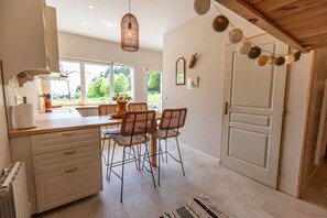 Une cuisine avec un coin repas, des meubles blancs, des chaises en osier, un luminaire suspendu et une vue par la fenêtre.