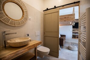 Une salle de bain moderne avec un grand miroir rond, un meuble-lavabo en bois, des toilettes blanches et une porte coulissante donnant sur un espace de vie doté d’un plafond en bois et d’un téléviseur.