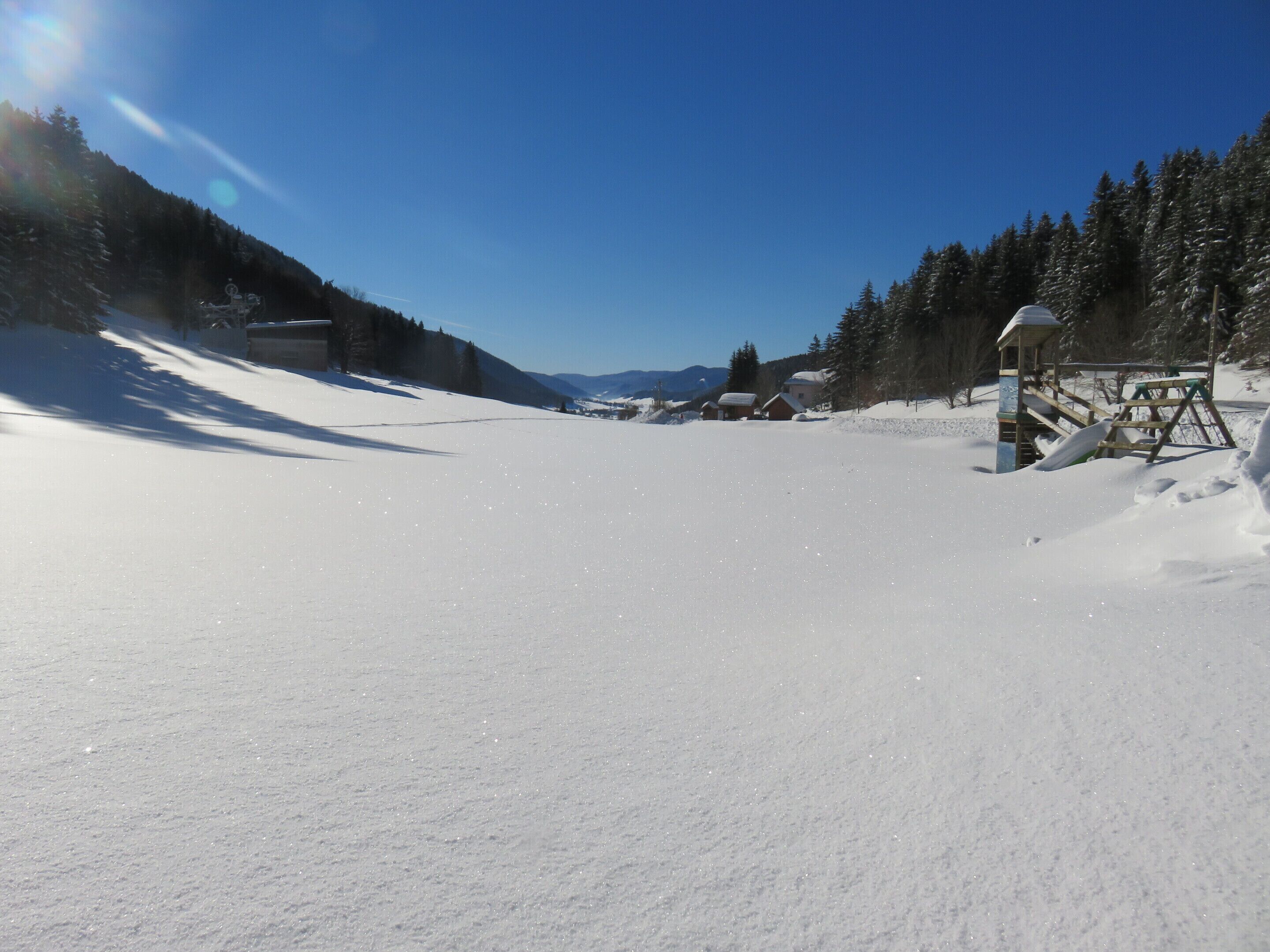 A snow-covered ski slope with a clear sky, a wooden ski lift, and buildings in the background.