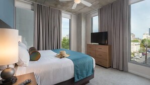 Ett hotellrum med en säng, en TV, en byrå och stora fönster med utsikt över staden.