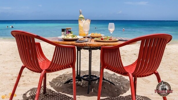 Ein Tisch am Strand mit zwei roten Stühlen, einer Flasche und einem Glas Wasser.