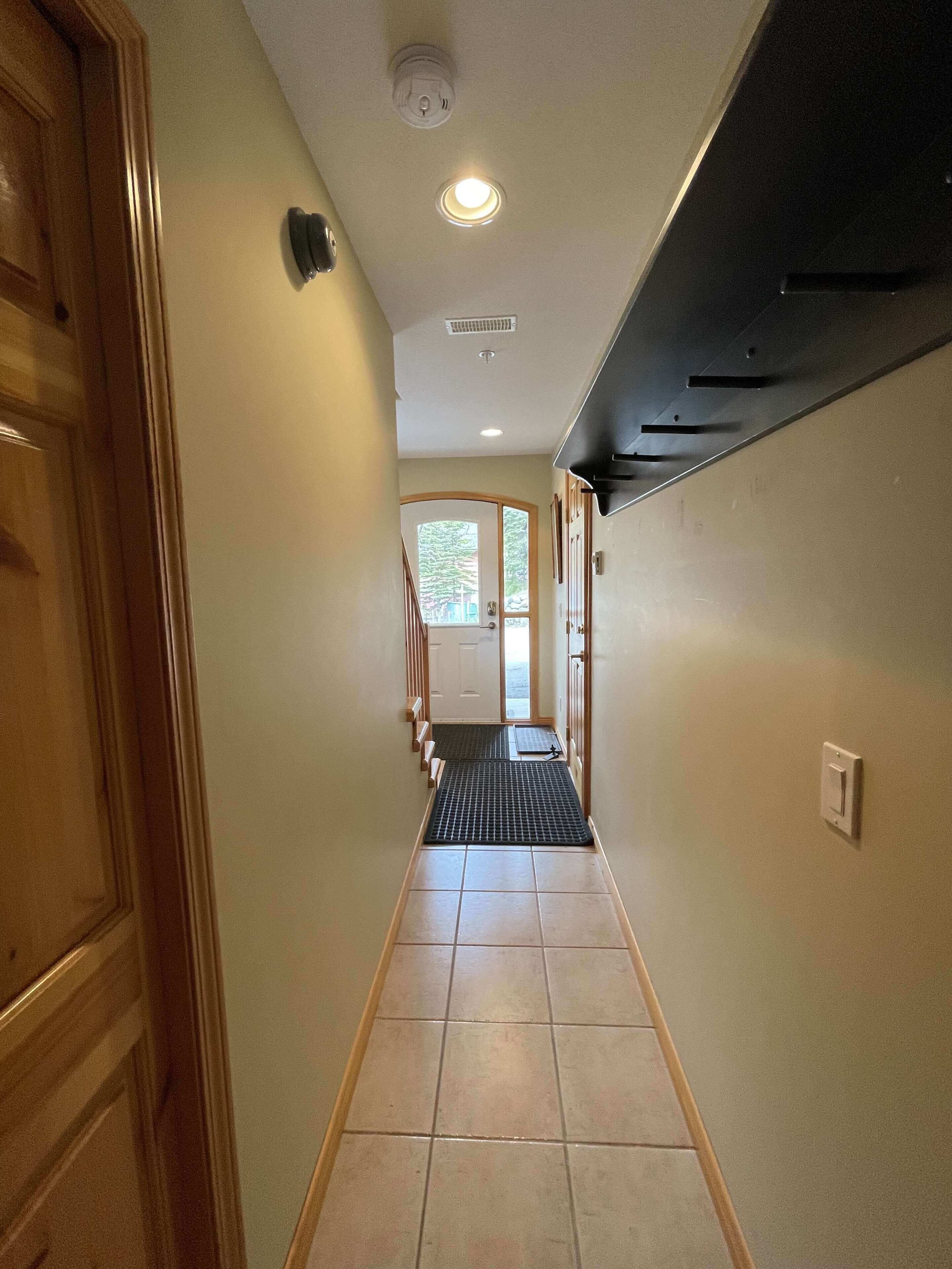 A narrow hallway with a wooden door on the left, a black metal shelf on the right, and a tiled floor.