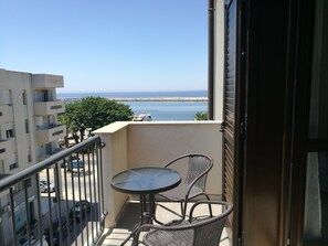 Un balcón con mesa y sillas, con vistas a una playa y un puerto con barcos.