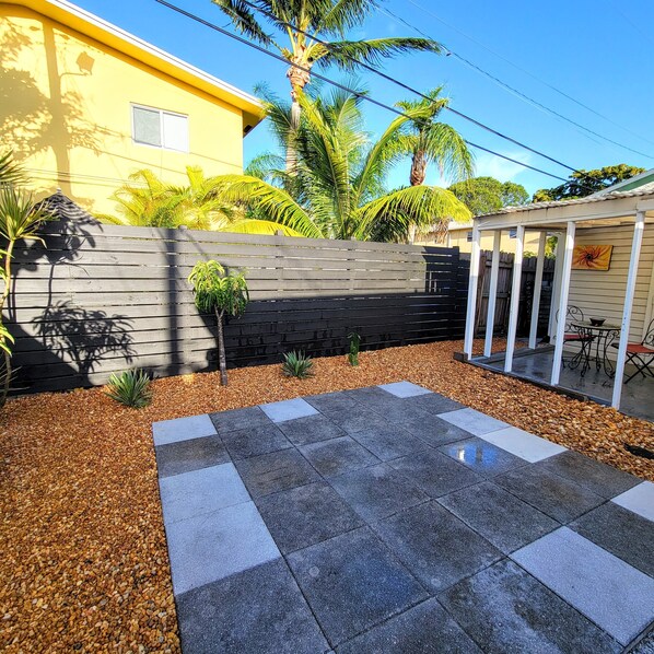 A patio with stone tiles and a wooden fence, surrounded by palm trees and a yellow building.