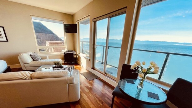 A spacious living room with a large sofa, a coffee table, and a view of the sea through large windows. - Points West Oceanfront Rooftop suite (Sooke)