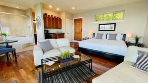A modern hotel room with a large bed, a sofa, a glass coffee table, and a view of the kitchen and outside. - Points West Oceanfront Rooftop suite (Sooke)
