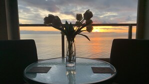 A round table with a vase of flowers and two placemats, positioned by a window overlooking a sunset over a body of water. - Points West Oceanfront Rooftop suite (Sooke)