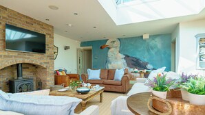 A cozy living room with a brick fireplace, comfortable seating, a large painting, and a dining area with a copper table. - This family home is situated in the gorgeous village of Bull Bay on Anglesey. (Bull Bay)