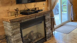 L’intérieur d’une cabane en rondins est agrémenté d’une cheminée en pierre, d’un téléviseur à écran plat fixé au mur et d’une porte-fenêtre coulissante donnant sur une terrasse.