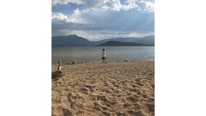 Eine Person steht auf einem Sandstrand, im Hintergrund Wasser und Berge.