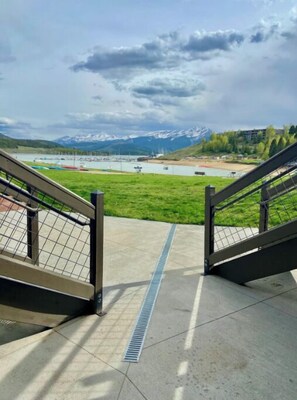 Vista desde un balcón con barandilla metálica, sobre un cuerpo de agua, una zona verde y montañas a lo lejos.