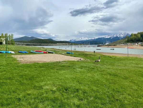 Un espacio verde con una red de voleibol, botes en un lago y montañas al fondo.