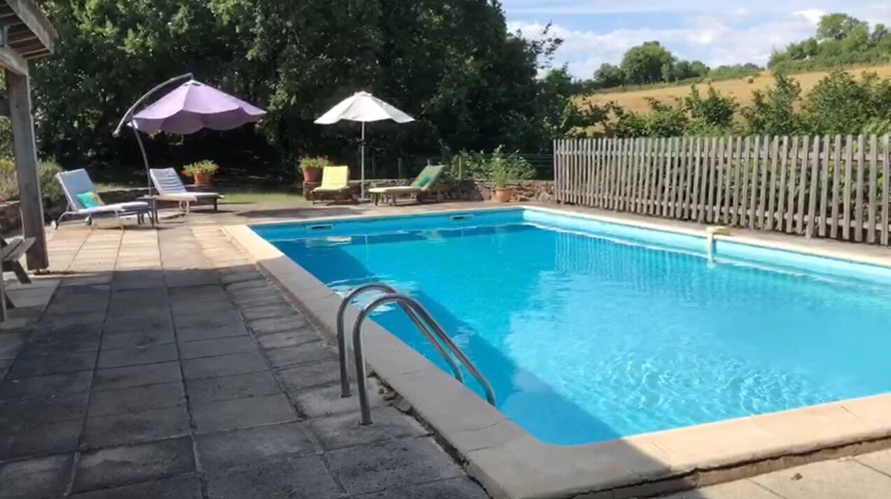 A rectangular swimming pool with a metal ladder on one side, surrounded by a tiled deck. There are lounge chairs and umbrellas in the background.