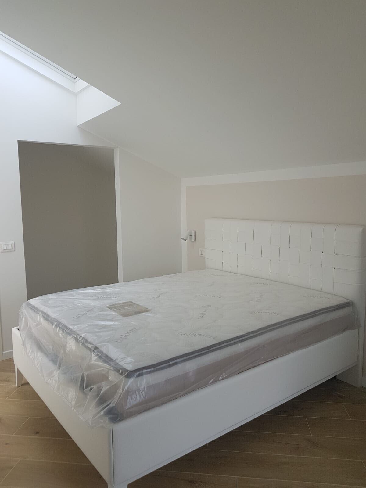 Un dormitorio con cama blanca, tragaluz en el techo inclinado y pared blanca lisa.