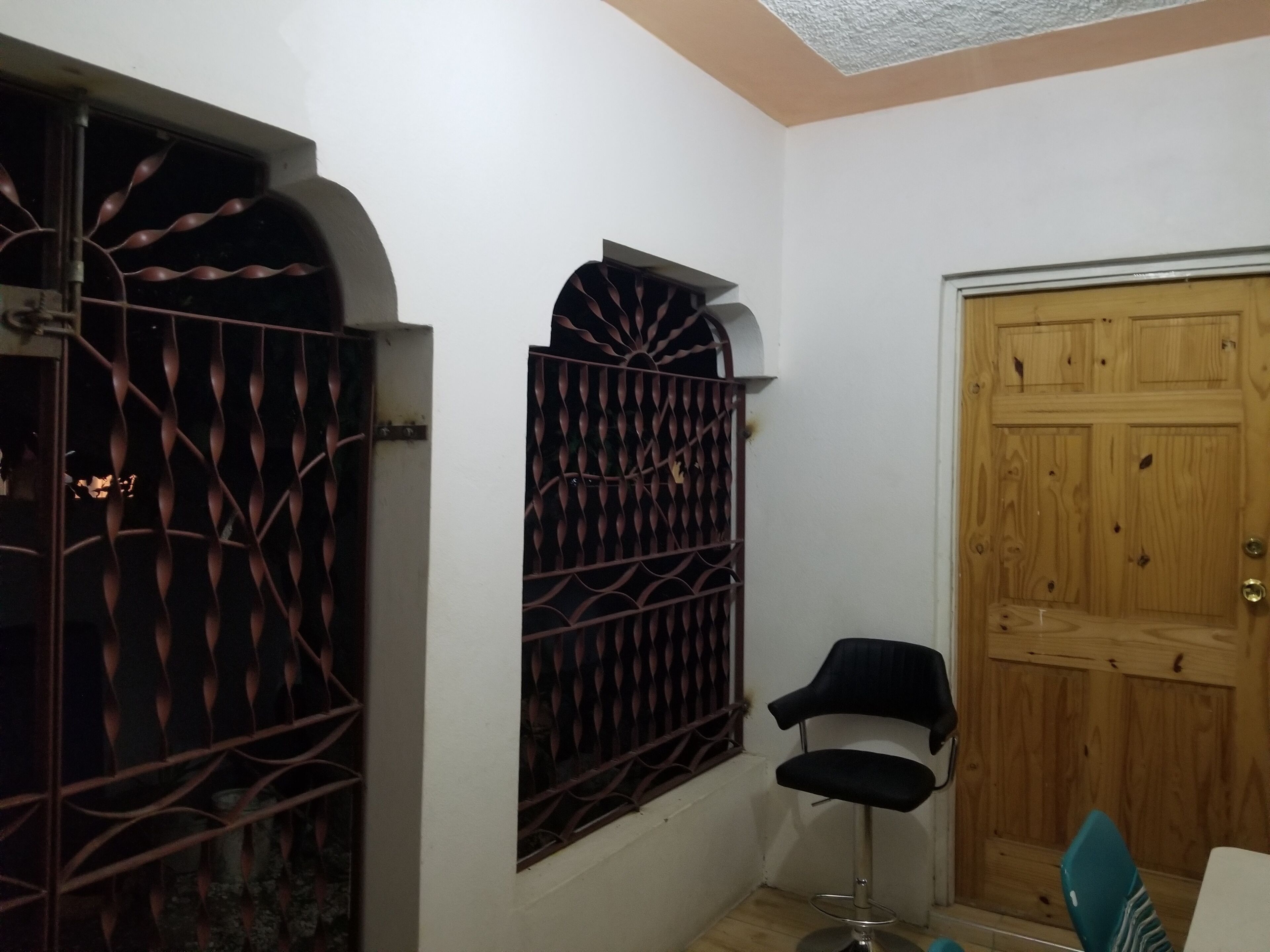 Una stanza con una porta in legno, una finestra con inferriata metallica e una sedia nera.