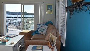 Un salón con vista a un puerto deportivo, un sofá con un cojín de lunares, un cuadro enmarcado en la pared y una mesita con un libro y objetos decorativos.