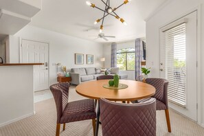 A modern living room with a round wooden dining table, two cushioned chairs, a ceiling fan, and a window with blinds.