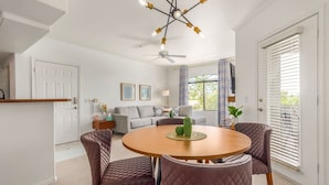 A modern living room with a round wooden table, two cushioned chairs, a ceiling fan, and a large window with blinds.