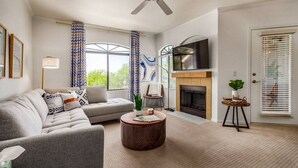 A living room with a sofa, a coffee table, a fireplace, a TV, and large windows with curtains.