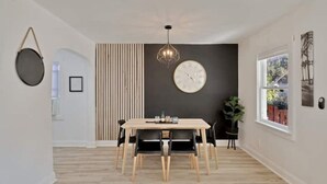 A dining area with a wooden table and chairs, a wall-mounted clock, and a window with a view of greenery. - ModernTropic 2bd/1ba Near Beach w/ fence yard/deck (Lake Worth)