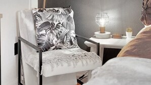 A modern room with a grey accent wall, a large green plant, a zebra-patterned pillow, a small table with a lamp, and a bed with a white cover.