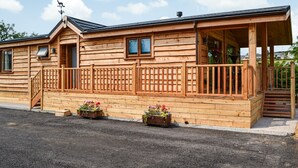 A wooden cabin with a porch, a chimney, and a small garden area with flowers. - Canadian timber lodge with direct views to the Alpacas. (Rowrah, near Cockermouth)