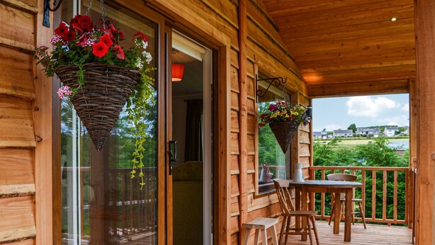 A wooden deck with a glass door leading to a room, a table with chairs, and hanging flower baskets. - Canadian timber lodge with direct views to the Alpacas. (Rowrah, near Cockermouth)