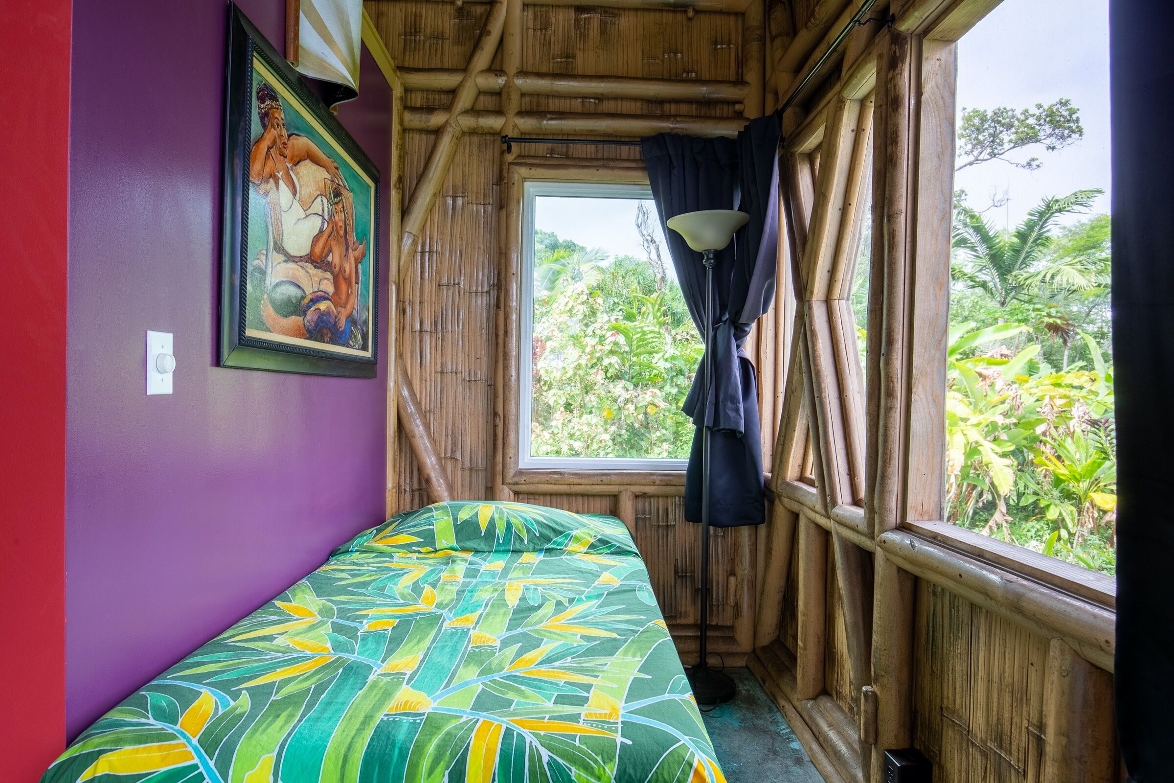 Une pièce avec un lit, une fenêtre avec des rideaux, un tableau au mur et une vue sur la verdure à l’extérieur.