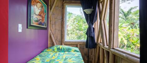 Une pièce avec un lit, une fenêtre avec des rideaux, un tableau au mur et une vue sur la verdure à l’extérieur.