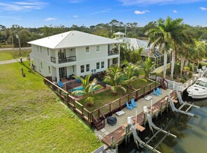 A two-story house with a deck overlooking a body of water, surrounded by palm trees and a dock.