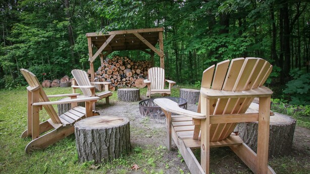 Un ensemble de chaises Adirondack en bois est disposé autour d’une table ronde en bois, lui-même entouré de souches d’arbres, dans une zone boisée où l’on aperçoit une pile de troncs sous une structure en bois.