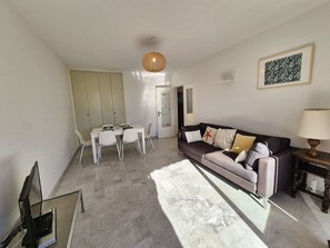 A modern living room with a sofa, a dining table, and chairs. There is a lamp on a side table and a framed picture on the wall.