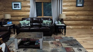 Ein Holzblockhaus-Interieur mit einem Ledersofa, zwei Nachttischen mit Lampen, einem Holztisch als Couchtisch und einem gemusterten Teppich.