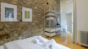 A bedroom with a stone wall, a bed with white linens, a white shelving unit, and a bathroom visible through an open door. - Incomparable Plaka's Lux 2bdrm Apt w/Pvt Heated Pool (Athens)