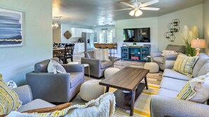 A modern living room with grey sofas, a wooden coffee table, and a TV. There is a dining area in the background with a chandelier and a kitchen visible through an open door. - Merritt Island Oasis w/ Pool ~ 7 Mi to Beach! (Merritt Island)