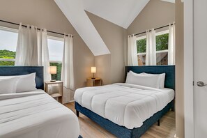 A bedroom with two beds, a nightstand, a lamp, and a view of greenery outside.