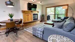 A modern living room with a grey sofa, wooden dining table, and chairs. There is a fireplace, a television mounted above it, and a large window providing natural light. - Targhee Creekside Condo - direct shuttle service to Targhee! (Driggs)