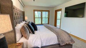 A bedroom with a large bed, a nightstand, a lamp, a TV, and a window with a view of trees. - Targhee Creekside Condo - direct shuttle service to Targhee! (Driggs)